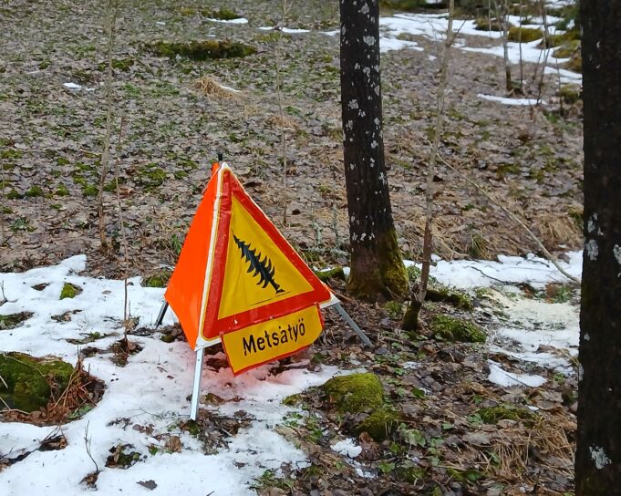 Kuvassa on vähälumisessa maastossa kelta-punainen varoituskyltti, jossa kaatuvan puun kuva ja teksti: Metsätyö.